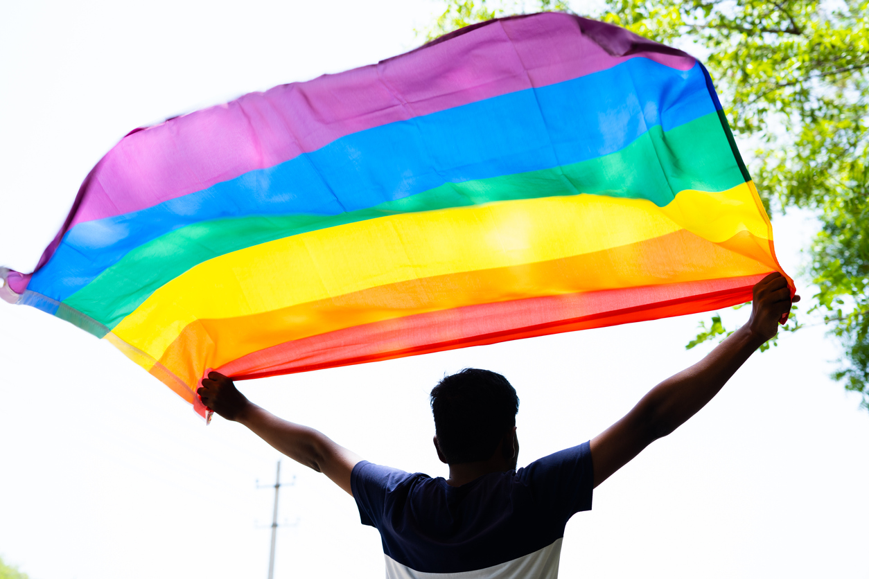Back view shot of young man proudly holding waving LGBTQ flag